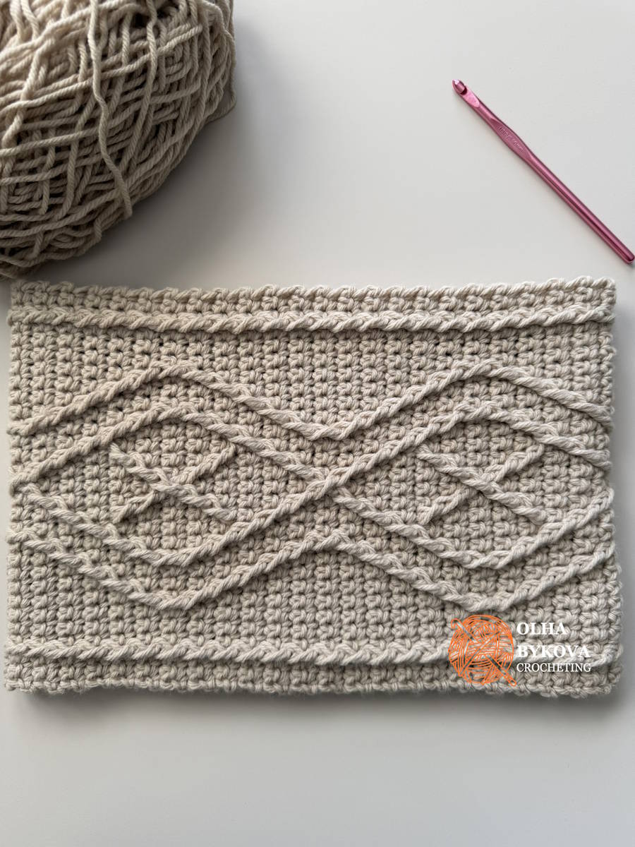 Diamond Frame crochet cable cowl laid flat beside worsted weight yarn and a crochet hook.